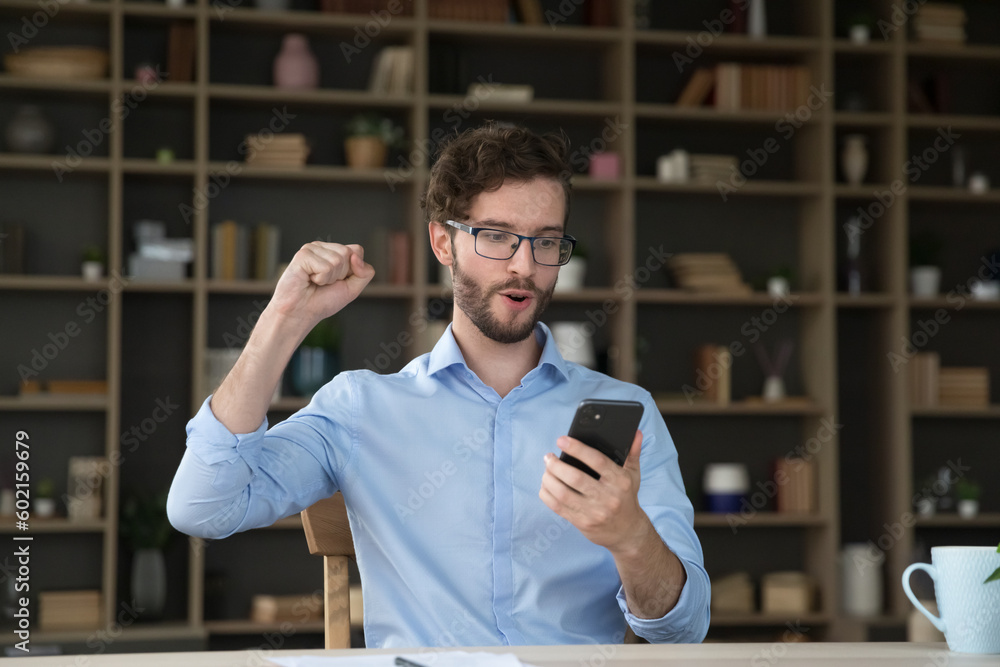 Surprised excited young business man holding mobile phone at home office table, making winner yes hand, victory gesture, shouting at screen, celebrating career success, win, job achievement Surprised excited young business man holding mobile phone at home office table, making winner yes hand, victory gesture, shouting at screen, celebrating career success, win, job achievement
