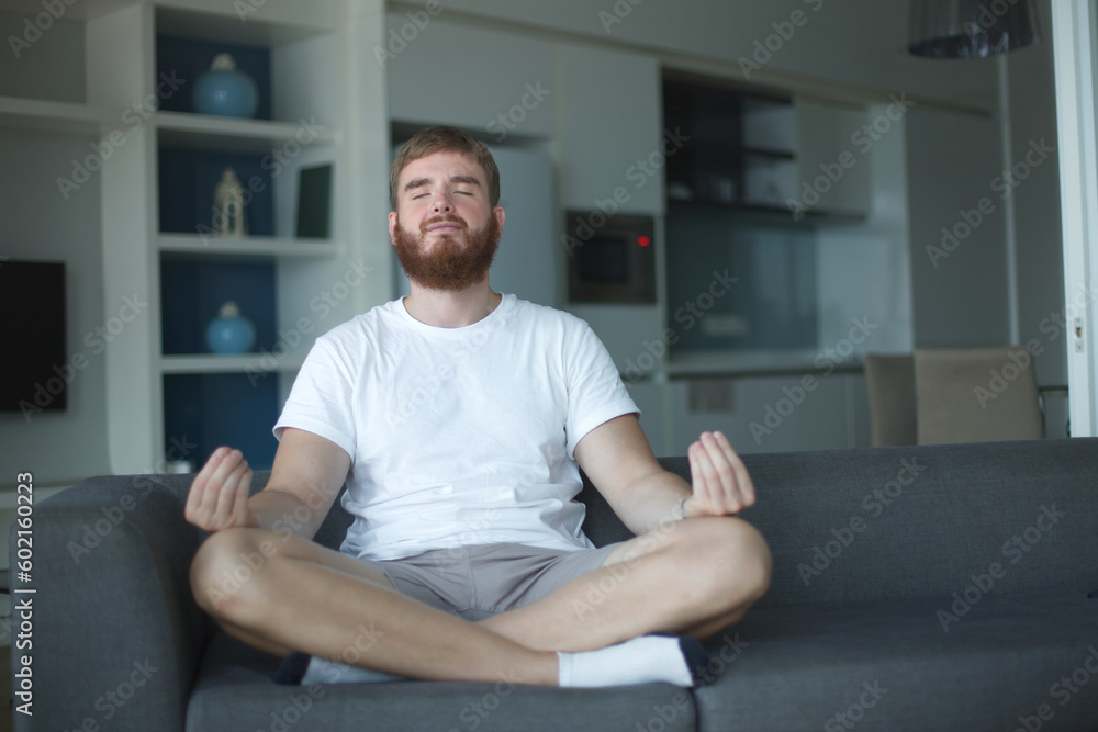 Young man with a beard alone at home, relax in living room ,sitting on the couch and meditating in the lotus position, in casual clothes Young man with a beard alone at home, relax in living room ,sitting on the couch and meditating in the lotus position, in casual clothes