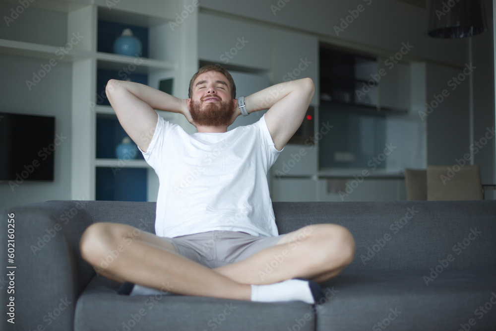 Young man with a beard alone at home, relax in living room ,sitting on the couch and meditating in the lotus position, in casual clothes Young man with a beard alone at home, relax in living room ,sitting on the couch and meditating in the lotus position, in casual clothes