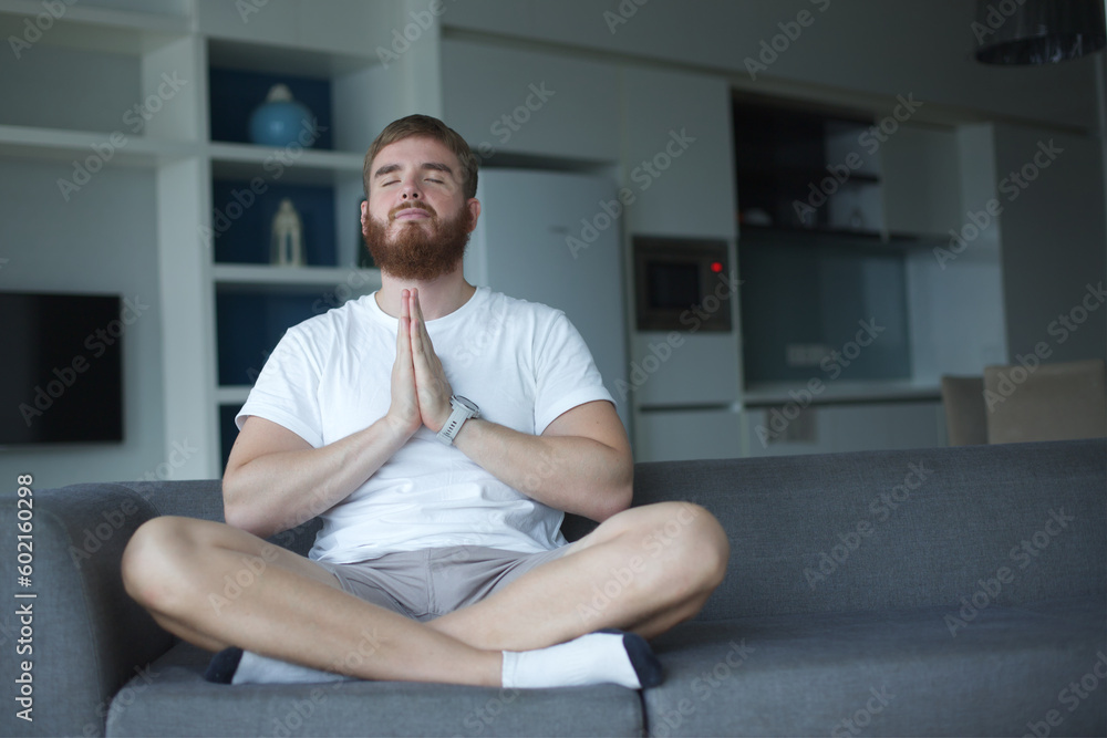Young man with a beard alone at home, relax in living room ,sitting on the couch and meditating in the lotus position, in casual clothes Young man with a beard alone at home, relax in living room ,sitting on the couch and meditating in the lotus position, in casual clothes