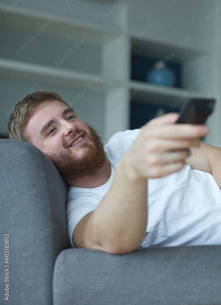 portrait of joyful young man in casual clothes sitting and lay on sofa in living room and looking tv with remote control in hand at home 