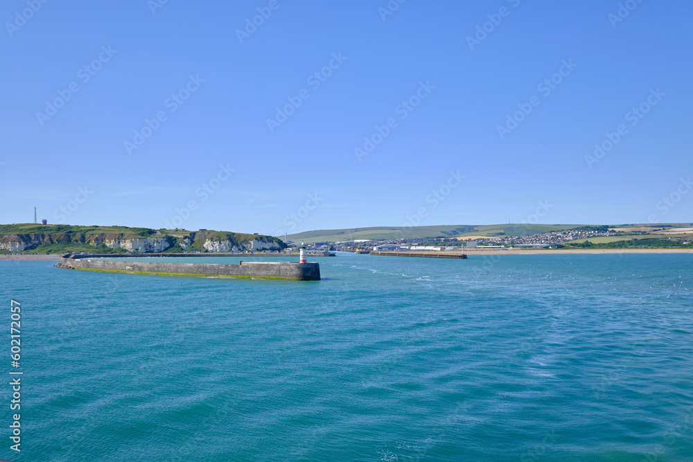 The entrance to Newhaven harbour, East Sussex, UK and its breakwater and lighthouse. Clear blue skiy and water with green hills in the background.