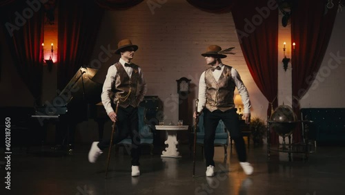 Two stylish professional male dancers in classy dandy hat performing jazz funk dance synchronic moves in vintage production studio retro environment with theatrical red velvet curtains and candlelight photo