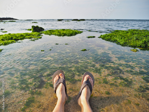 A person wearing flip-flops, in the afternoon, the rocks with seaweed on the seaside, leisurely stretching his feet at Qianshui Bay in Sanzhi District, Taiwan.