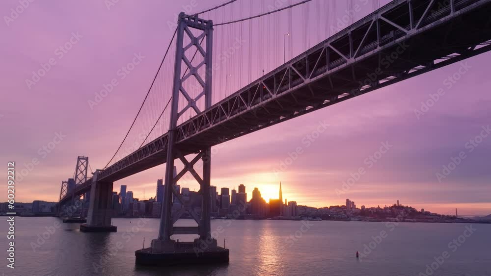 Drone flying above water surface under Bay Bridge construction with scenic cityscape silhouette on background. Beautiful aerial skyscrapers in downtown San Francisco in cinematic pink cloudy sky 4K Drone flying above water surface under Bay Bridge construction with scenic cityscape silhouette on background. Beautiful aerial skyscrapers in downtown San Francisco in cinematic pink cloudy sky 4K