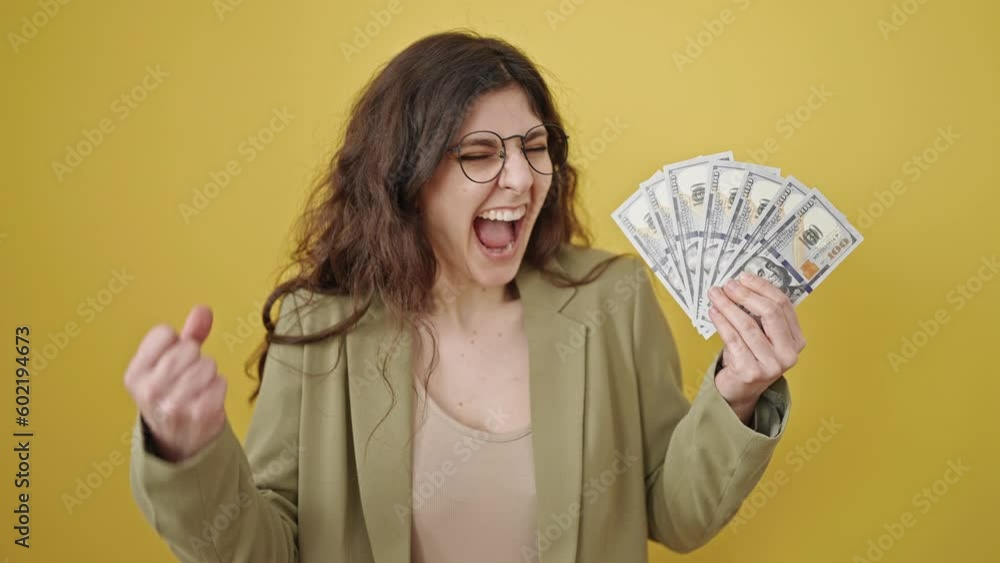 Young beautiful hispanic woman holding dollars with winner expression over isolated yellow background Young beautiful hispanic woman holding dollars with winner expression over isolated yellow background