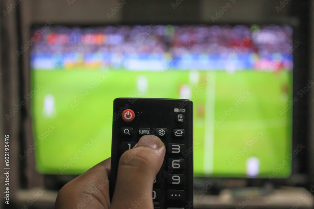 A man is pointing a black remote control at an unfocused tv while watching football A man is pointing a black remote control at an unfocused tv while watching football