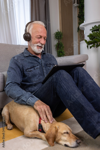 Senior man sits on the floor with his dog and using tablet pc. Elderly man with headphones listening to music, podcast, watch movie, video call with family or friends. Leisure and retirement concept