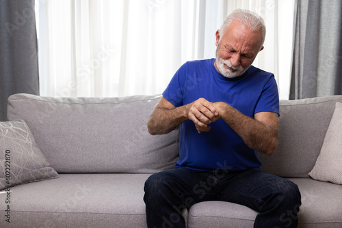 Elderly man holding his wrist, suffering from arthritis pain in hand, sitting on sofa at home © Space_Cat