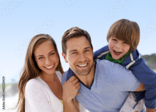 Beach, family and summer portrait outdoor for travel, adventure or holiday and piggy back fun. A man, woman and child or son playing together on vacation at sea with a blue sky, parents and happiness