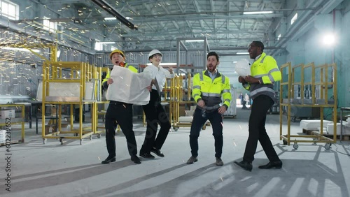 Multicultural engineers dancing at modern manufacturing factory and clapping hands. Employees in safety hard hats celebrating succsess at background of welding sparks flying. photo