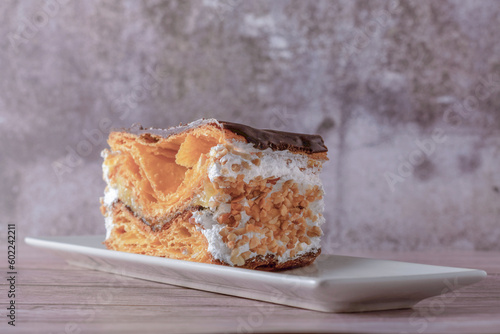 close-up of a mille-feuille filled with cream and almonds covered with chocolate on a white plate with ceramic spoons with chocolate and cocoa powder