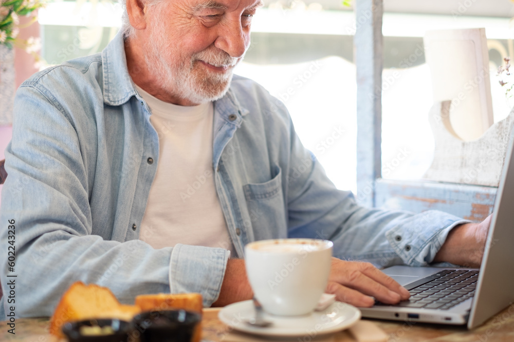 Smiling senior man in coffee shop having breakfast with cappuccino and sweet cake while working on laptop. Elderly people enjoying free lifestyle and pleasant moment of relax Smiling senior man in coffee shop having breakfast with cappuccino and sweet cake while working on laptop. Elderly people enjoying free lifestyle and pleasant moment of relax