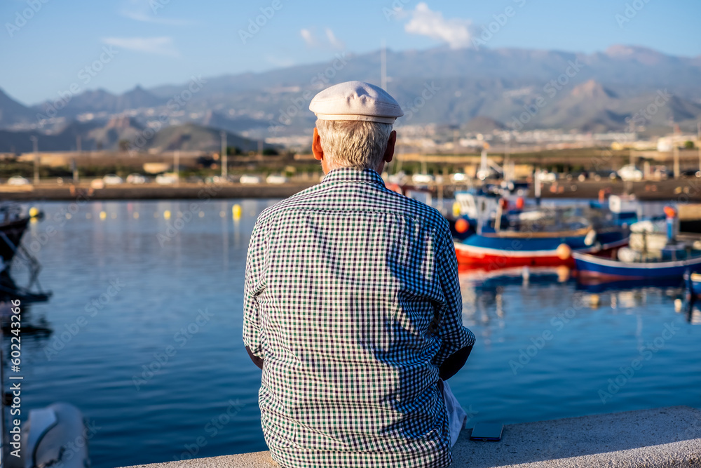 Back view of senior man in casual shirt and hat sitting at the port looking at boats and  sea. Relaxed elderly bearded male enjoying travel or retirement and free time