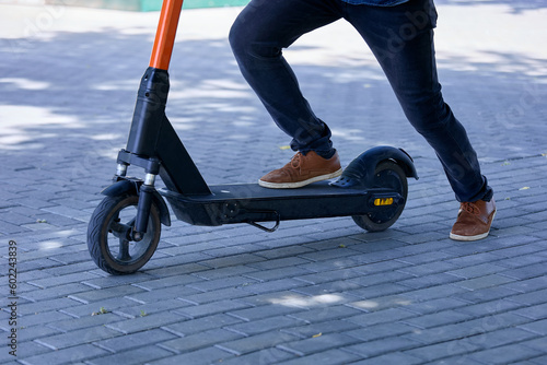 A young man walks through the park on an electric scooter along the sidewalks. Close-up of a guy in fashionable sneakers pushes off with his foot to ride an electric scooter