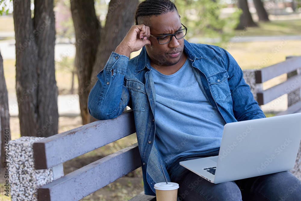 Fototapeta premium Modern businessman carefully checks reports of employees sitting on bench on summer day. Stylish man in denim shirt performs work in comfortable place outside the office thanks to modern technology