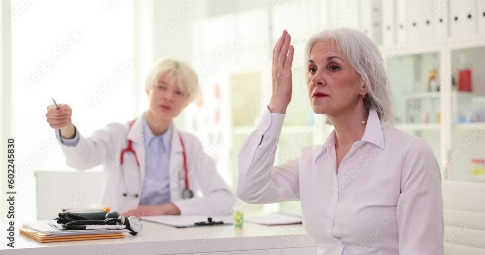 Female ophthalmologist points pen to letters on Snellen chart. Mature woman closes eye with hand while sitting on chair in specialist office slow motion Female ophthalmologist points pen to letters on Snellen chart. Mature woman closes eye with hand while sitting on chair in specialist office slow motion