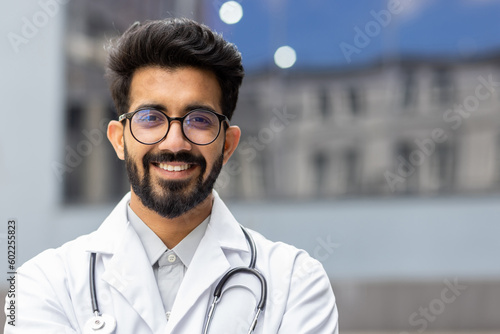 Close-up portrait of Hindu doctor, man in glasses smiling and looking at camera, doctor student intern with stethoscope outside clinic.