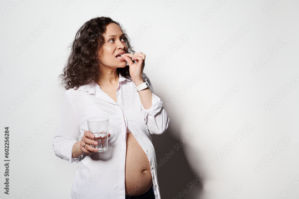 Multi-ethnic adult pregnant woman holding glass of water and pill. Concept of pregnancy, health care, gynecology, medicine and healthy lifestyle. Mother expecting her baby. Nutritional supplements. Multi-ethnic adult pregnant woman holding glass of water and pill. Concept of pregnancy, health care, gynecology, medicine and healthy lifestyle. Mother expecting her baby. Nutritional supplements.