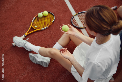 Top view of young girl in sportswear sitting on tennis court while holding ball in hand. © Davidovici