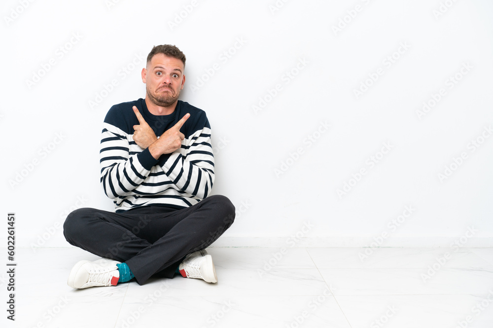 Young man sitting on the floor isolated on white background pointing to the laterals having doubts