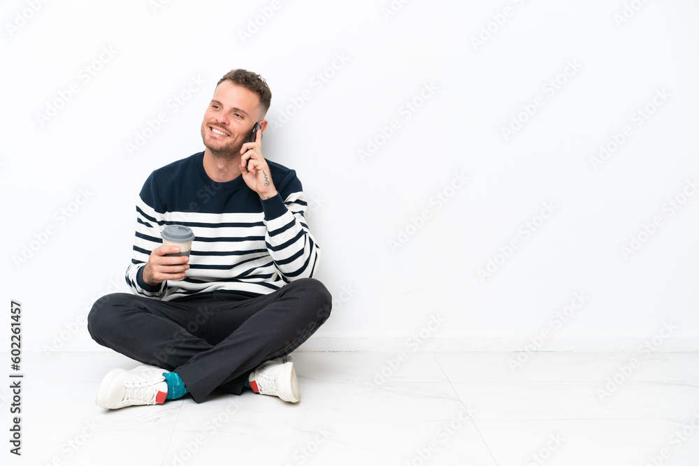Young man sitting on the floor isolated on white background holding coffee to take away and a mobile Young man sitting on the floor isolated on white background holding coffee to take away and a mobile