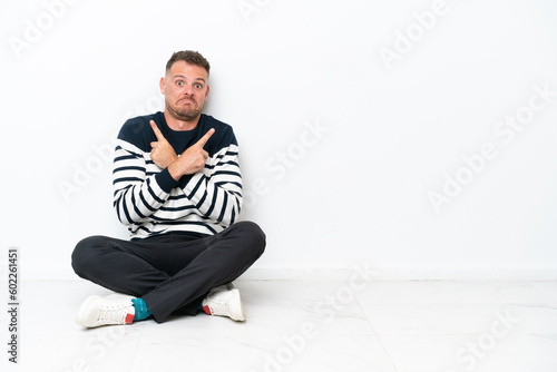 Young man sitting on the floor isolated on white background pointing to the laterals having doubts