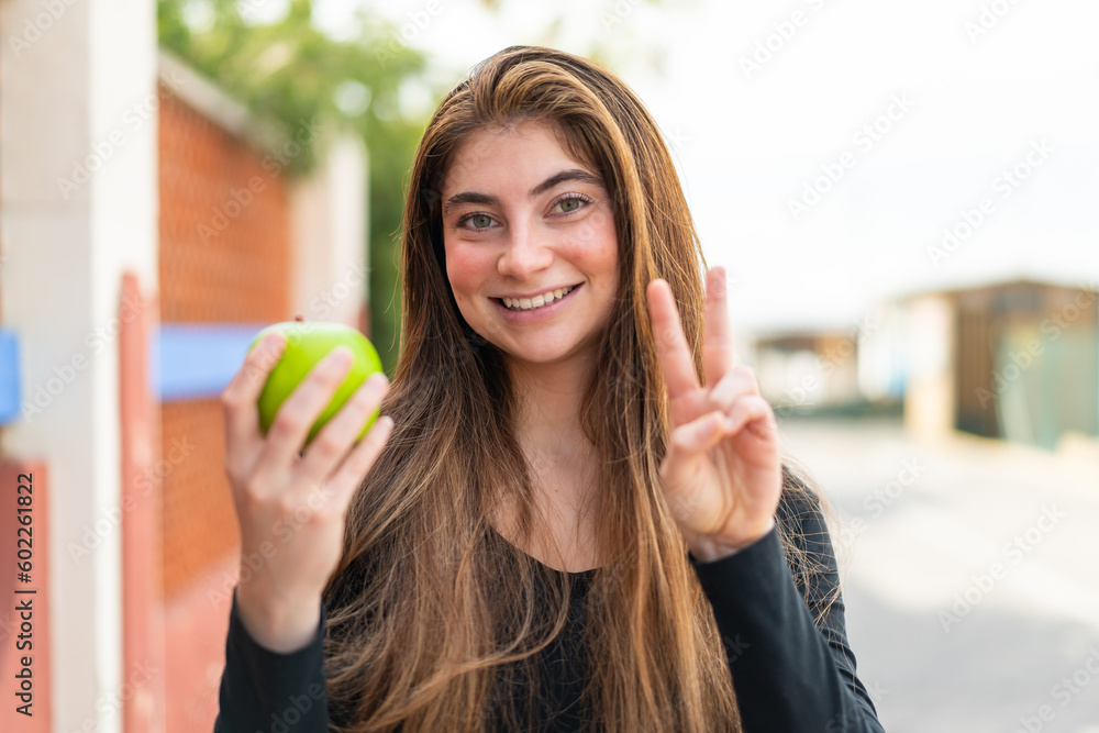 Young pretty caucasian woman with an apple at outdoors smiling and showing victory sign Young pretty caucasian woman with an apple at outdoors smiling and showing victory sign