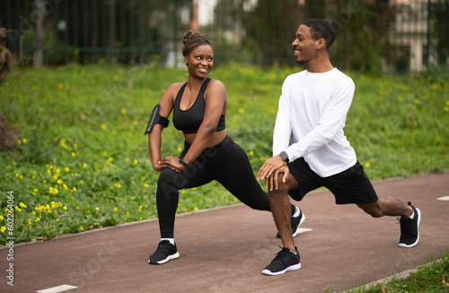 Positive young african american couple in sportswear doing leg exercise, stretching and warm up in park