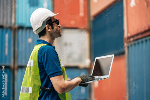 Engineer dock worker in protective safety jumpsuit uniform and with hardhat and use laptop computer at cargo container shipping warehouse. transportation import,export logistic industrial