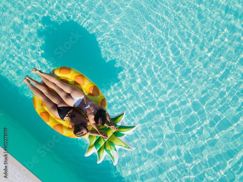 Top view of two girls relaxing on inflatable big pineapple in the pool. Aerial view of ladies relaxing on the floating mattress.