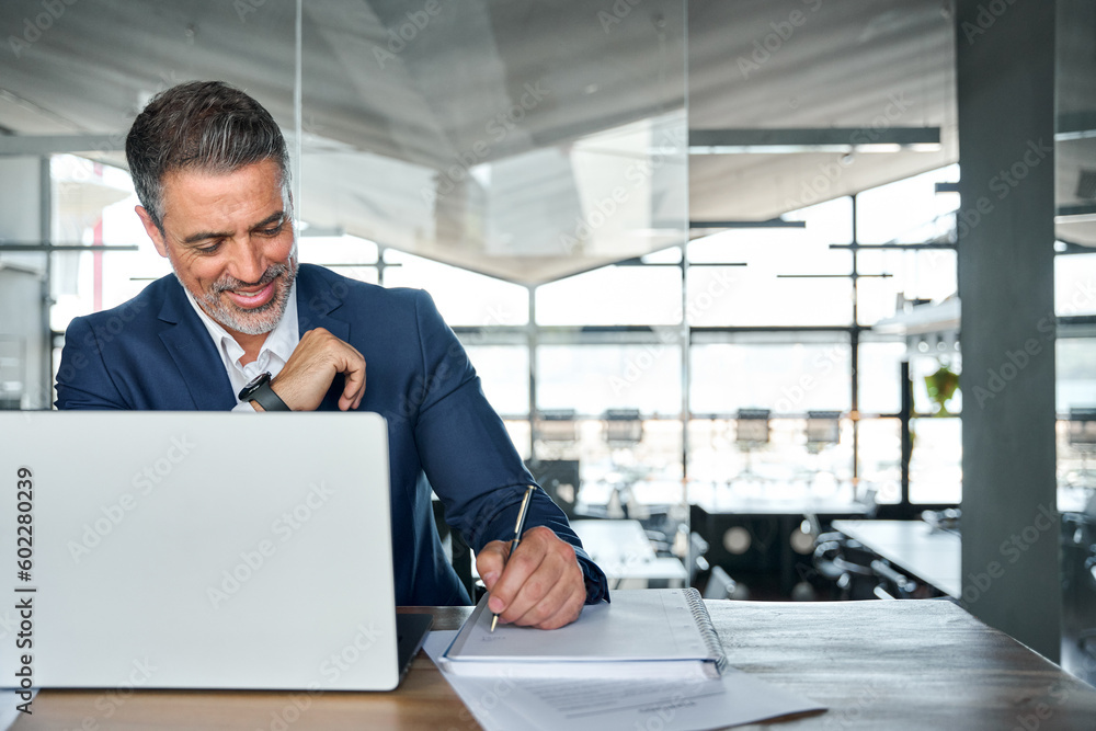 Busy happy middle aged smiling professional business man company executive ceo manager or lawyer wearing suit sitting at desk in modern office working on laptop computer writing notes, copy space. Busy happy middle aged smiling professional business man company executive ceo manager or lawyer wearing suit sitting at desk in modern office working on laptop computer writing notes, copy space.