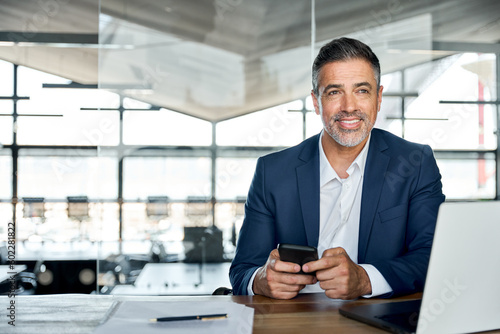 Smiling mid aged business man ceo wearing blue suit sitting in office using cell phone solutions. Mature businessman professional executive holding mobile working at desk with laptop and smartphone.