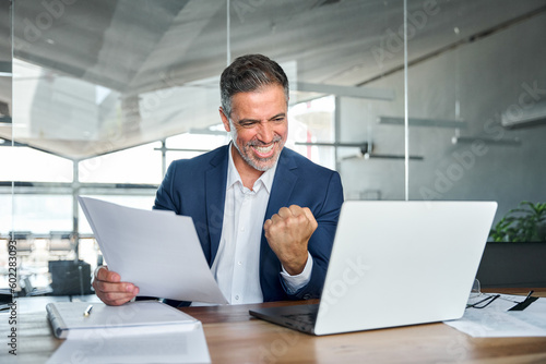 Happy mature older business man ceo wearing suit celebrating success at work in office holding papers looking at laptop rejoicing company growth, goals achievement good results screaming yes.