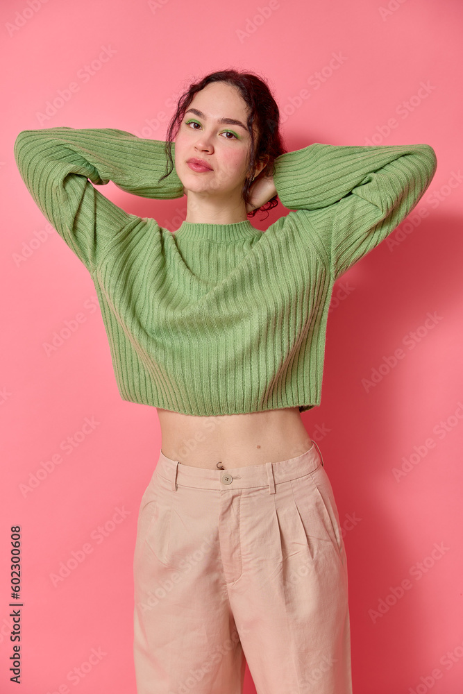 Φωτογραφία Vertical shot of beautiful slim young woman with dark curly hair vivid make up green eye shadow stands with hands behind head wears green cropped sweater and pants shows bare belly on pink background