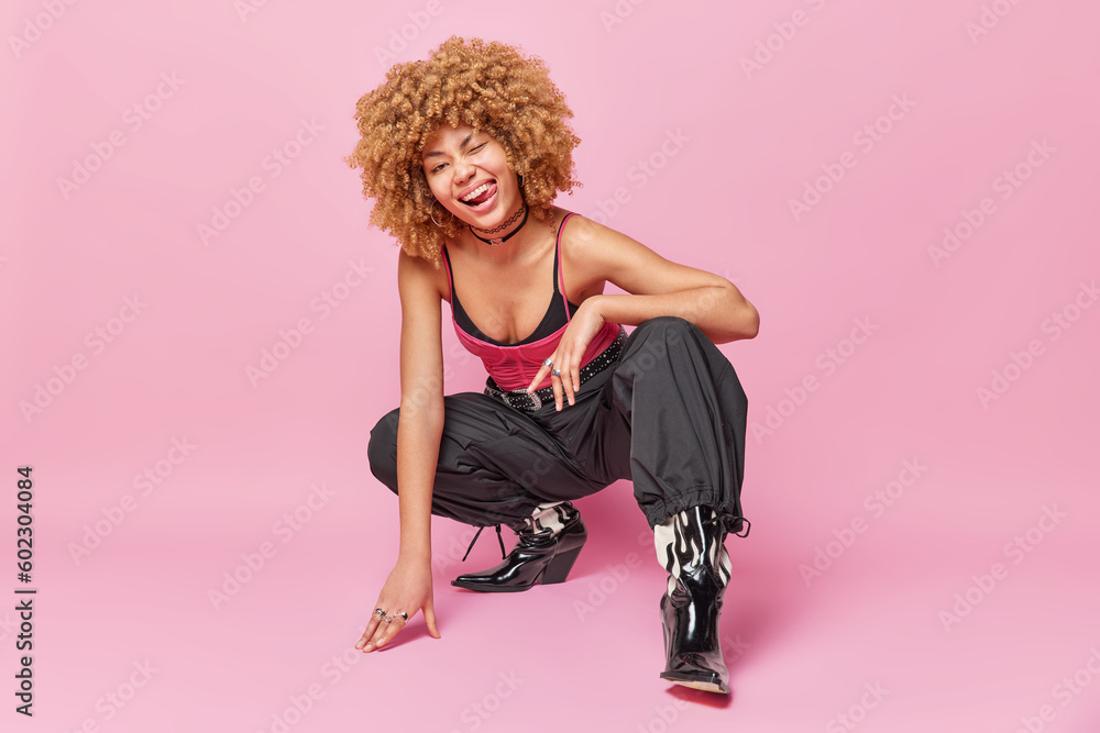 Full length shot of fashionable curly woman squats against pink background winks eye and sticks out tongue wears stylish clothing isolated over pink backgroun has flirty expression. Fashion model - obrazy, fototapety, plakaty 