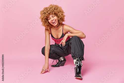 Full length shot of fashionable curly woman squats against pink background winks eye and sticks out tongue wears stylish clothing isolated over pink backgroun has flirty expression. Fashion model