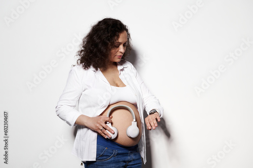 Pregnant multi ethnic woman, expectant mom placing headphone on her bare belly, turning on cool music for her future newborn baby isolated on white background. Maternity and happy carefree pregnancy