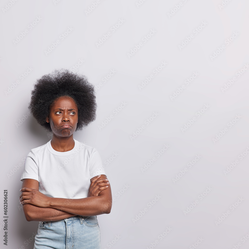 Vertical shot of displeased African woman with curly hair stands in thoughtful expression keeps arms folded concentrated aside has discontent expression dressed in t shirt and jeans white background Vertical shot of displeased African woman with curly hair stands in thoughtful expression keeps arms folded concentrated aside has discontent expression dressed in t shirt and jeans white background