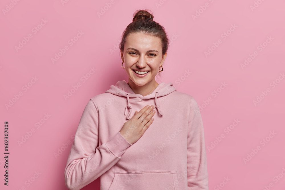 Horizontal shot of sincere happy woman laughs pleasantly keeps hand on chest giggles positively at something very funny dressed in sweatshirt isolated on pink background. People and authentic emotions Horizontal shot of sincere happy woman laughs pleasantly keeps hand on chest giggles positively at something very funny dressed in sweatshirt isolated on pink background. People and authentic emotions
