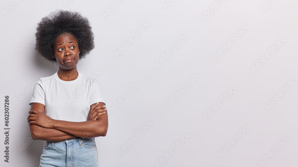 Horizontal shot of doubtful dark skinned woman keeps arms folded looks aside with curiosity thinks over interesting offer wears casual t shirt jeans isolated on white background with blank copy space