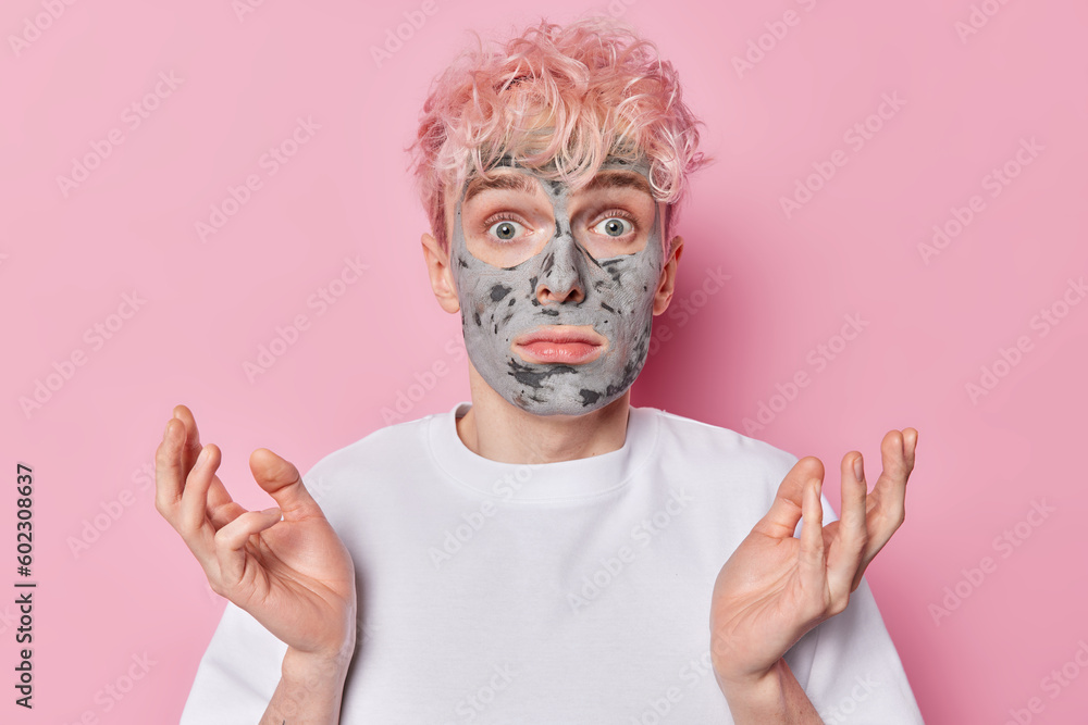 Clueless adult man spreads palms looks surprisingly at camera applies homemade clay mask on face to unclog pores stands hesitant indoor dressed in white t shirt isolated over pink background Clueless adult man spreads palms looks surprisingly at camera applies homemade clay mask on face to unclog pores stands hesitant indoor dressed in white t shirt isolated over pink background