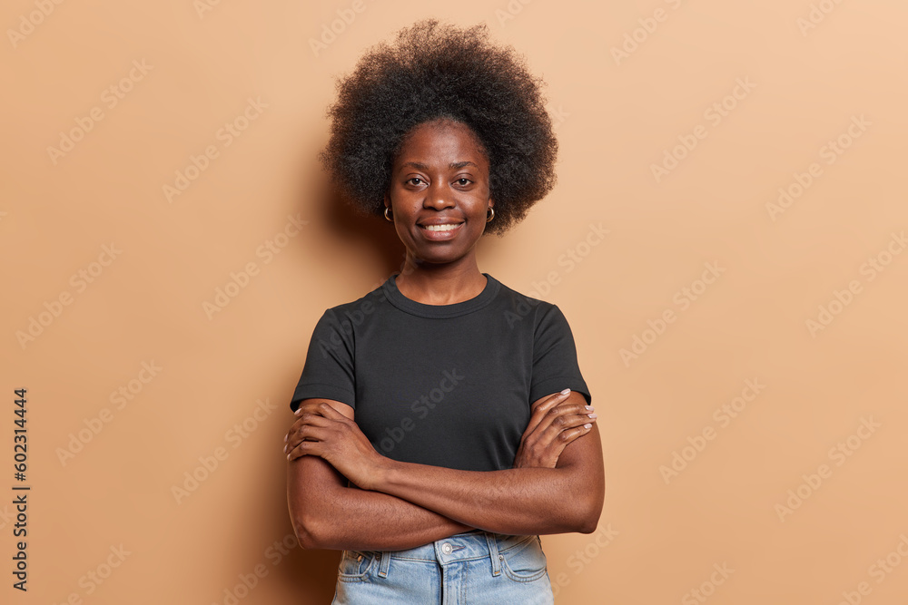 Self confident pretty young African woman with bushy hair with arms folded smiles gently dressed in casual black t shirt and jeans isolated over brown studio background feels assured. Indoor shot. Self confident pretty young African woman with bushy hair with arms folded smiles gently dressed in casual black t shirt and jeans isolated over brown studio background feels assured. Indoor shot.