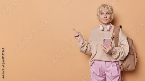 Studio shot of fair haired young woman wears pullover and pink trousers holds smartphone points index finger at copy space poses with rucsack advertises something isolated over brown background photo
