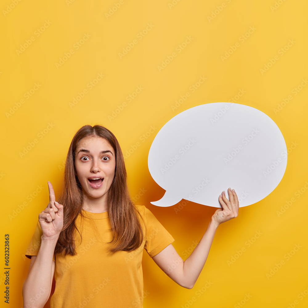 Impressed dark haired woman has idea or answer on question keeps index finger raised up holds blank speech bubble for your advertising content dressed in casual t shirt isolated over yellow background - obrazy, fototapety, plakaty 