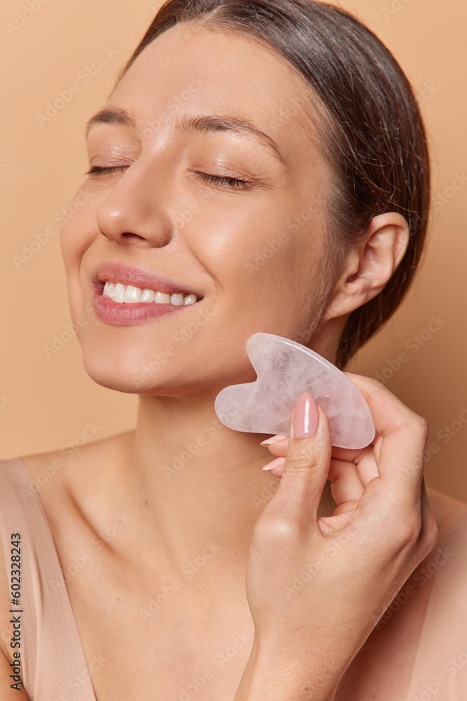 Headshot of dark haired woman enjoys guasha face massage with jade stone undergoes spa procedures at home for glowing skin gets energy and blood moving poses indoor. Self care and pampering concept