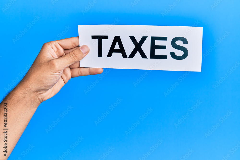 Hand of caucasian man holding paper with taxes word over isolated blue background Hand of caucasian man holding paper with taxes word over isolated blue background