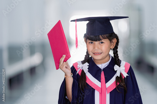 A cheerful kid graduate wearing an academic gown and graduation cap celebrates her graduation in the classroom