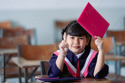 A cheerful kid graduate wearing an academic gown and graduation cap celebrates her graduation in the classroom