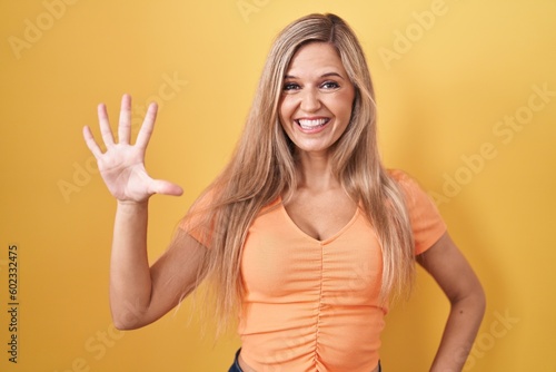 Young woman standing over yellow background showing and pointing up with fingers number five while smiling confident and happy.